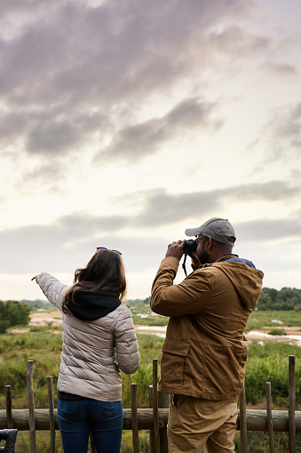 Rear view of a young couple using binoculars to look at the scenic view of a wildlife reserve from an observation deck during a safari