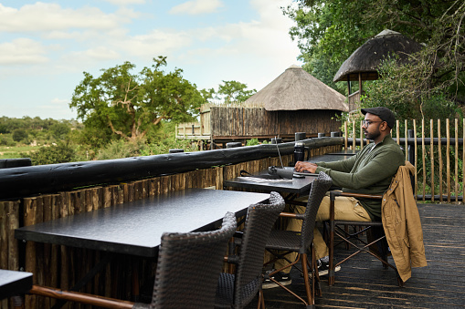 Young man sitting at a table and working on a laptop in the restaurant of a wildlife reserve with a scenic view