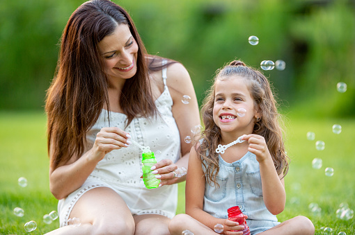 A sweet little girl sits with her mother in the grass on a warm summers day as they play with bubble wands and blow them into the wind.  They are each dressed casually and are smiling as they enjoy the fresh air and each others company.