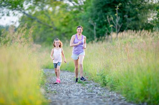 A mother and her young daughter take a run together outside on a sunny summer day.  They are both dressed comfortably in athletic wear as they spend time exercising in the fresh air together.