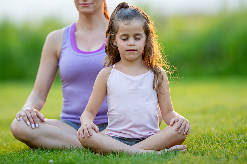 A mother and her young daughter sit outside in the grass cross-legged as they meditate together.  They are both dressed comfortably in athletic wear as they close their eyes and focus on their breathing as they take in the fresh air.