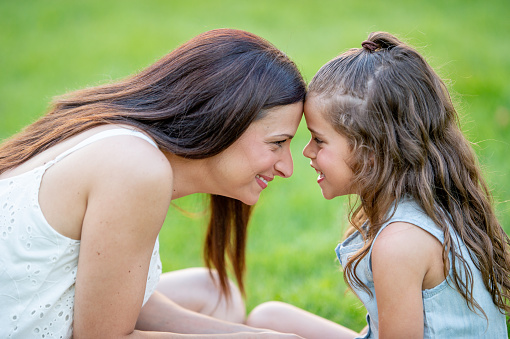 A young mother and her daughter sit face-to-face with their foreheads touching as they pose for a portrait.  They are both dressed casually and smiling as they enjoy the fresh air together.