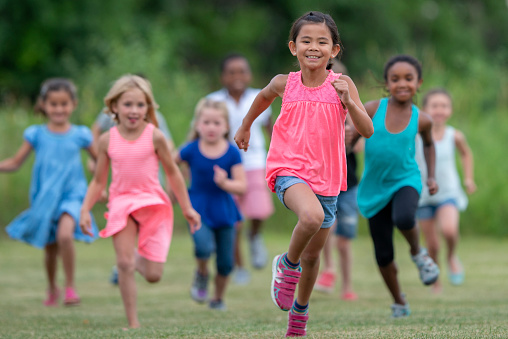A large group of Elementary aged children are seen running out side together on a summer day.  They are each dressed casually and are smiling as they play together.