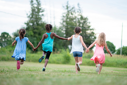A small group of four school aged children, hold hands as they run through the grass together on a sunny summer day.