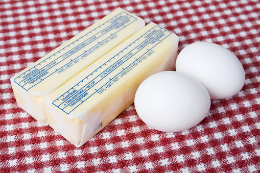 Two sticks of packaged butter and two eggs on a red and white checkered tablecloth.