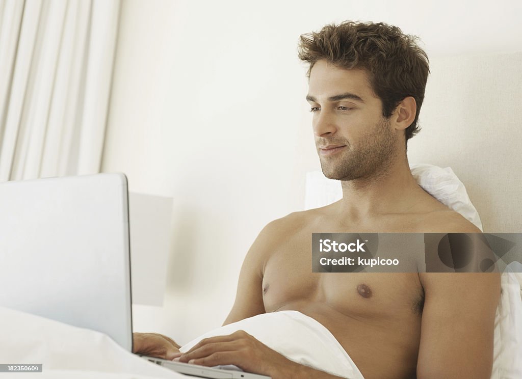 Hombre atractivo joven usando una computadora portátil en la habitación mientras está en la cama - Foto de stock de 20 a 29 años libre de derechos