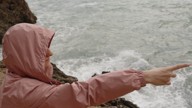 A Woman Stands Among the Rocks by the Sea in Strong Wind, Wearing a Pink Hooded Raincoat, and Points Somewhere with Her Finger.