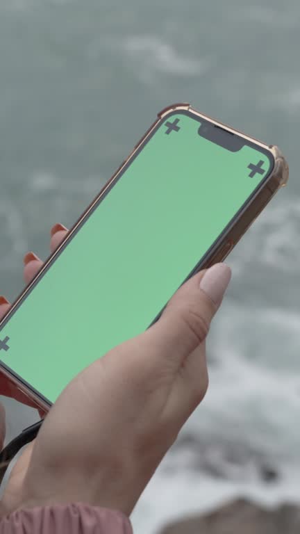 Vertical video. Smartphone with a Green Screen and Tracking Markers is Held by a Woman with Long Hair Standing on the Edge of a Cliff by the Sea. Overcast Weather and Large Waves.