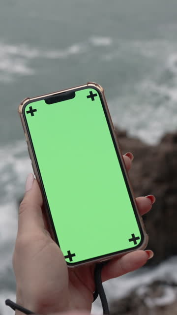 Vertical video. View From Behind the Female Head as She Holds a Smartphone with a Green Screen While Standing on the Edge of a Cliff by the Stormy Sea with Large Waves and Overcast Weather.