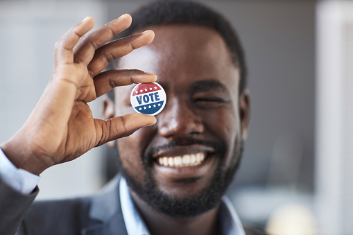 Focus on hand of young smiling African American man holding vote badge in front of his right eye while standing in front of camera