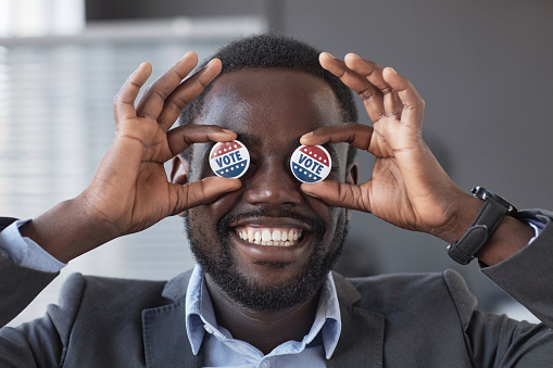 Happy young African American voter with wide toothy smile holding two small round vote badges by his eyes while standing in front of camera