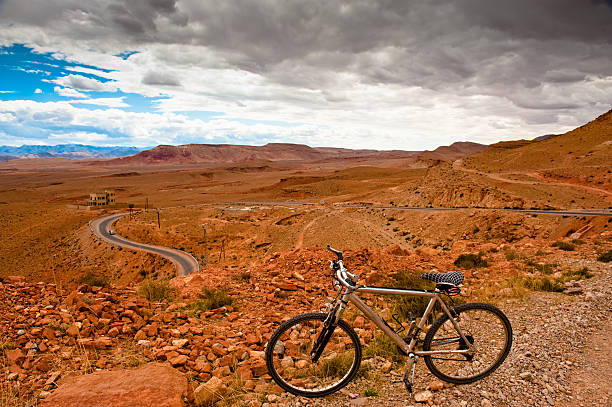 Distant view of a cyclist riding a dirt track in the Patagonian steppe. The massive, jagged granite peaks of Mount Fitz Roy loom in the background under a clear blue sky in Argentina. Epic scale.