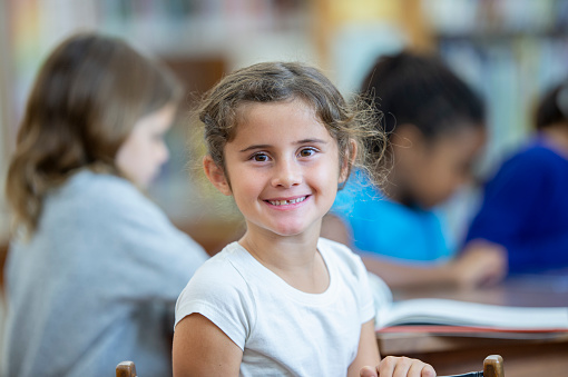 A young elementary student of mixed race, sits at a table in the library as she reads independently.  She has turned around in her chair to smile for the camera.