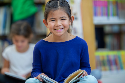 A young elementary student of Asian decent, sits cross legged on the floor in the library as she reads independently.  She is dressed casually and is looking up from her book to smile.