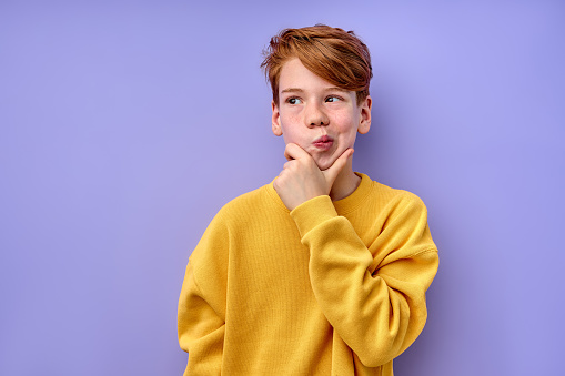 Let me think. Caucasian pensive clever redhead schooler in yellow shirt with puzzled serious expression, child thinking doubting, making choice. indoor studio shot isolated on purple background