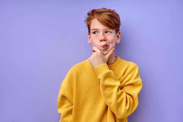 clever redhead schooler with puzzled serious expression thinking, looking at side Let me think. Caucasian pensive clever redhead schooler in yellow shirt with puzzled serious expression, child thinking doubting, making choice. indoor studio shot isolated on purple background critical thinking kids stock pictures, royalty-free photos & images