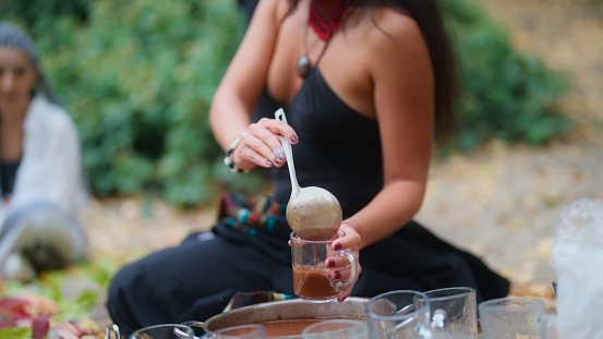 A woman in a black dress is serving cacao drinks during a cacao ceremony in nature. \nA cacao ceremony is a spiritual ritual for cleaning soul, connecting with the nature, healing, mental wellbeing, mindfulness and self-exploration. People who join the ceremony consume ceremonial cacao.