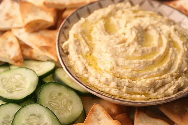 High-resolution, full-frame, close-up digital capture of a bowl of freshly made hummus with sliced Arabian cucumbers and golden toasted pita points. Surface of hummus is drizzled with extra virgin olive oil.