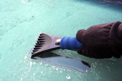 A driver wearing gloves scraping the ice off his windshield in the morning.