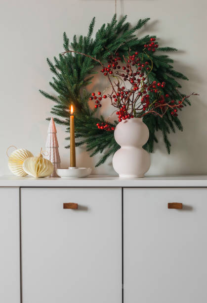 istock Christmas tree wreath on the wall, a vase with cranberry branches, a burning candle, Christmas decor on a white chest of drawers 1823857413
