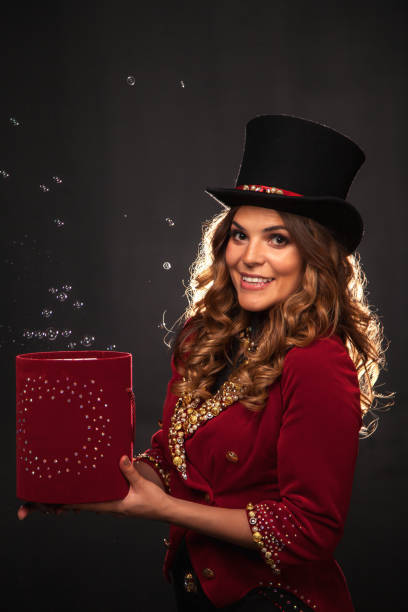 istock Circus actress lady in top hat holding magic red box at stage shadow, looking at camera. Magician illusionist in theatrical costume shows soap bubbles show. Theatre perform concept. Copy ad text space 1823990960