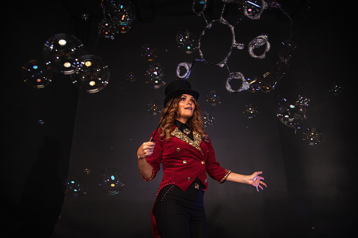 Magician illusionist woman in red costume performing soap bubbles show, happy look. Chic circus actor lady in top hat showing at shadow theatre stage. Theatrical perform concept. Copy ad text space