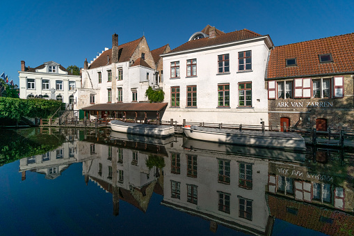Bruges, Belgium - July 07, 2023: Wharf on the canal since Dijver park with the Our Lady church tower reflected on the canal in the old town of the beautiful city of Brugge in Belgium in a sunny day.