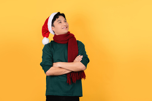 Handsome young Asian man in green Christmas outfit, arms crossed and looking up on yellow background.