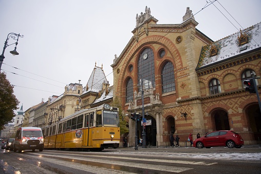 Budapest, Hungary - November 30, 20203: Central Market Hall. Pedestrian crossing and yellow tram in foreground, snowy old building in background.