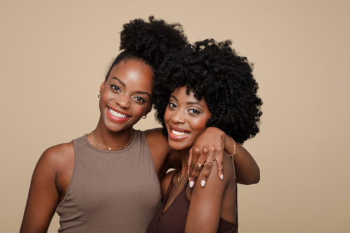 Two beautiful young women wearing brown clothes standing against beige background, embracing and smiling.