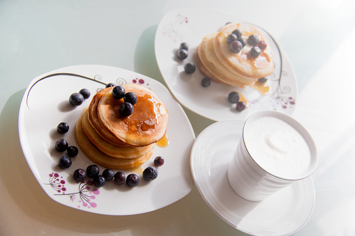 Homemade pancake plates topped by maple syrup, cinnamon and blueberries on white plates with cup of delicious coffee topped with cream, yummy and sweet breakfast table for two