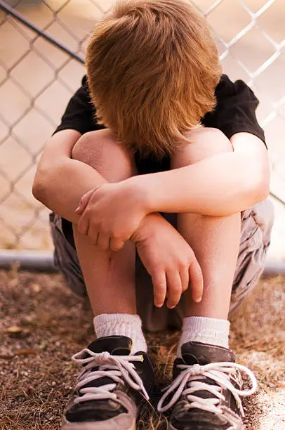 A unhappy little boy is leaning against a chain-link fence.