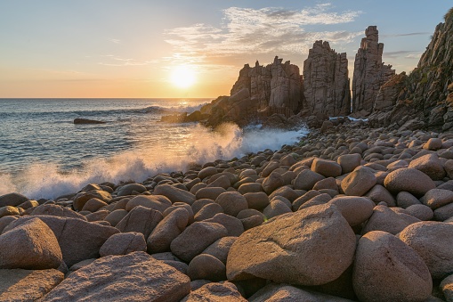 A beach shore with large rocks and crashing waves illuminated by the warm glow of an orange sky