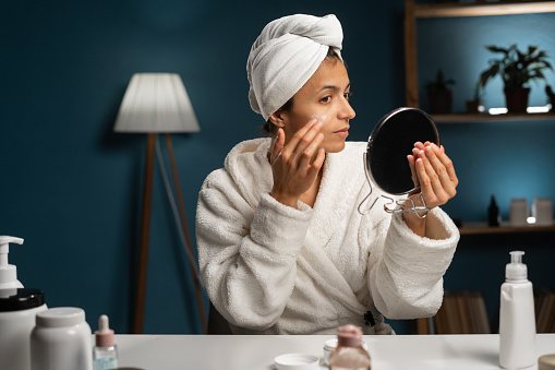Beautiful hispanic woman applying face cream sitting at the table. Smiling young woman in bathrobe doing skin care routine at home. Copy space