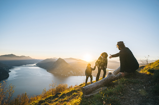 Father offers helping hand to son and mother on mountain top above lake at sunset