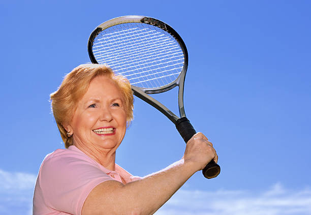 une femme âgée souriante jouant tennis contre un ciel bleu - senior adult outdoors wellbeing sky photos et images de collection