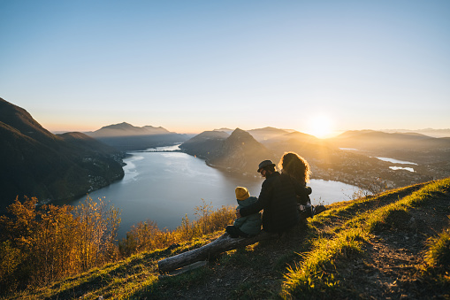 Family sits on mountain top together and looks out to lake and sunset