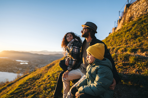 Family sits on mountain top together and looks out to lake and sunset