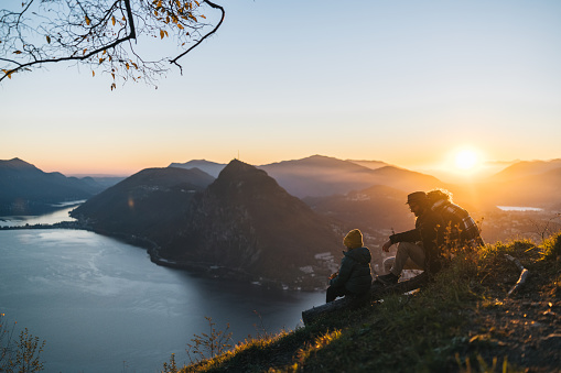 Family sits on mountain top together and looks out to lake and sunset