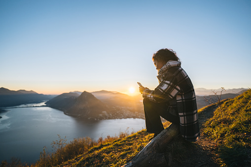 Young woman uses mobile phone on mountain top above lake at sunset