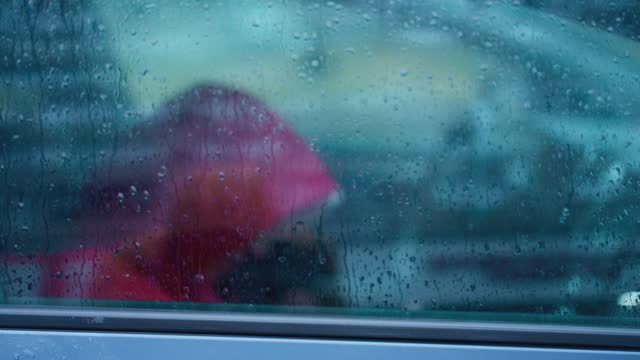silhouette of person in red jacket with hoodie sitting in car under autumn rain in city. shooting through window with water drops. heavy rainy season, cold wet fall.