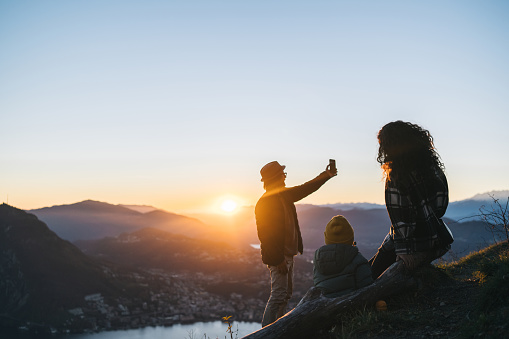 Family takes photo of son and mother on mountain top above lake at sunset