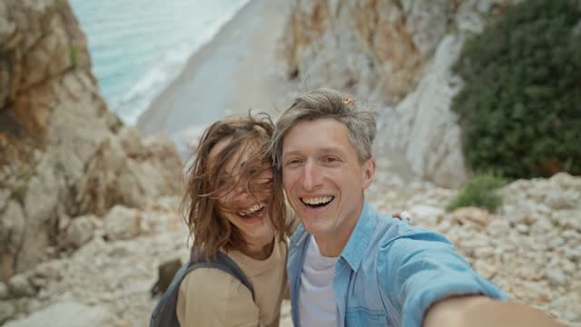 happy lovely couple young smiling woman and mid aged man hiking rocky canyon at seaside and taking selfie. wanderlust and travel summer vacation trip. using phone camera for filming.