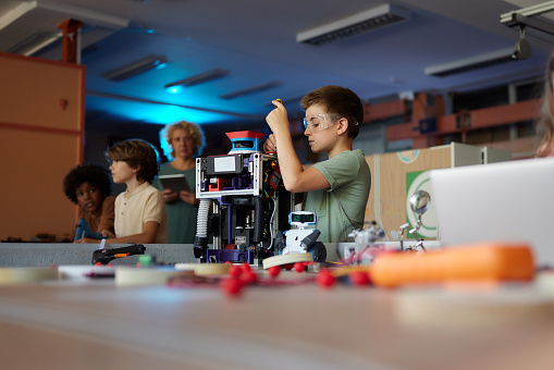 Little boy making an innovative robot during a class with his classmates and a teacher in a lab. Copy space.