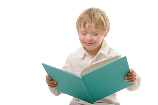 Seven year old boy with Down Syndrome dressed in white clothing reads a blue book. White background.