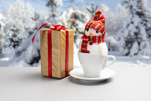 Gift box, a white cup with a toy snowman and a winter forest on the background. Christmastime celebration.