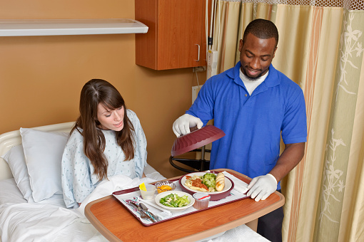 An aide serves a patient a meal in a hospital room.