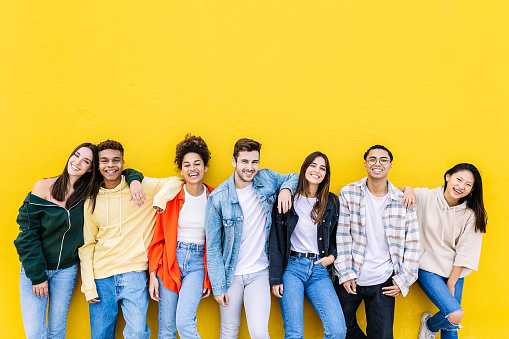 Young adult united group of multiracial happy friends leaning on yellow wall - Community and unity concept with diverse trendy teenage people smiling at camera