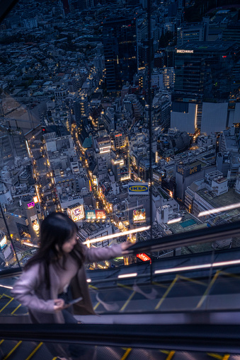 This photo captures a poignant moment of a young woman standing on the observation elevator of the Shibuya Sky building, as she gazes out over the dazzling nightscape of Shibuya district in Tokyo, Japan. The viewer is drawn into the scene from her perspective, sharing the awe that the panoramic city view inspires. The sprawling urban landscape below is a network of illuminated streets, vibrant signage, and dense buildings—a showcase of Shibuya's pulsating life after dark. The composition beautifully contrasts the solitary figure with the complex tapestry of city lights, suggesting a narrative of personal reflection amidst the urban rush.
