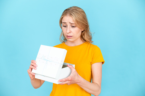 Portrait of sad displeased woman standing with opened gift package isolated on blue background. Wrong present on christmas. Negative human emotion, facial expressions, feelings, attitude reaction